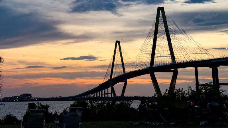 People relaxing on the lawn at the Mount Pleasant waterfront at dusk, with the Ravenel Bridge beyond